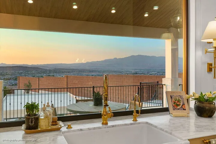 Expansive window overlooking distant mountains and a residential area, featuring a white undermount sink, gold-tone faucet, and stone-finish countertop