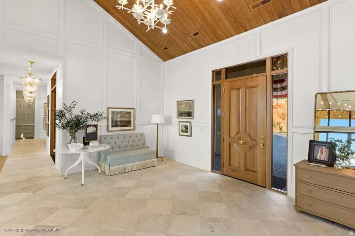 Vaulted wood-paneled ceiling, decorative wall molding, light-toned tile flooring, solid wood entry door with sidelights, and an ornate chandelier