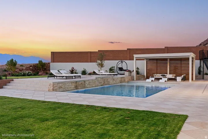In-ground swimming pool with a surrounding paved deck, stone-faced retaining wall, and a covered patio structure