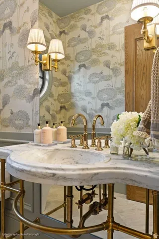 Marble vanity with integrated basin, brass fixtures, detailed patterned wallpaper, and a circular window