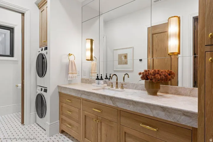 Wood-finish vanity with gold-tone hardware, stacked washer and dryer, full-wall mirrored installation, and dual wall sconces