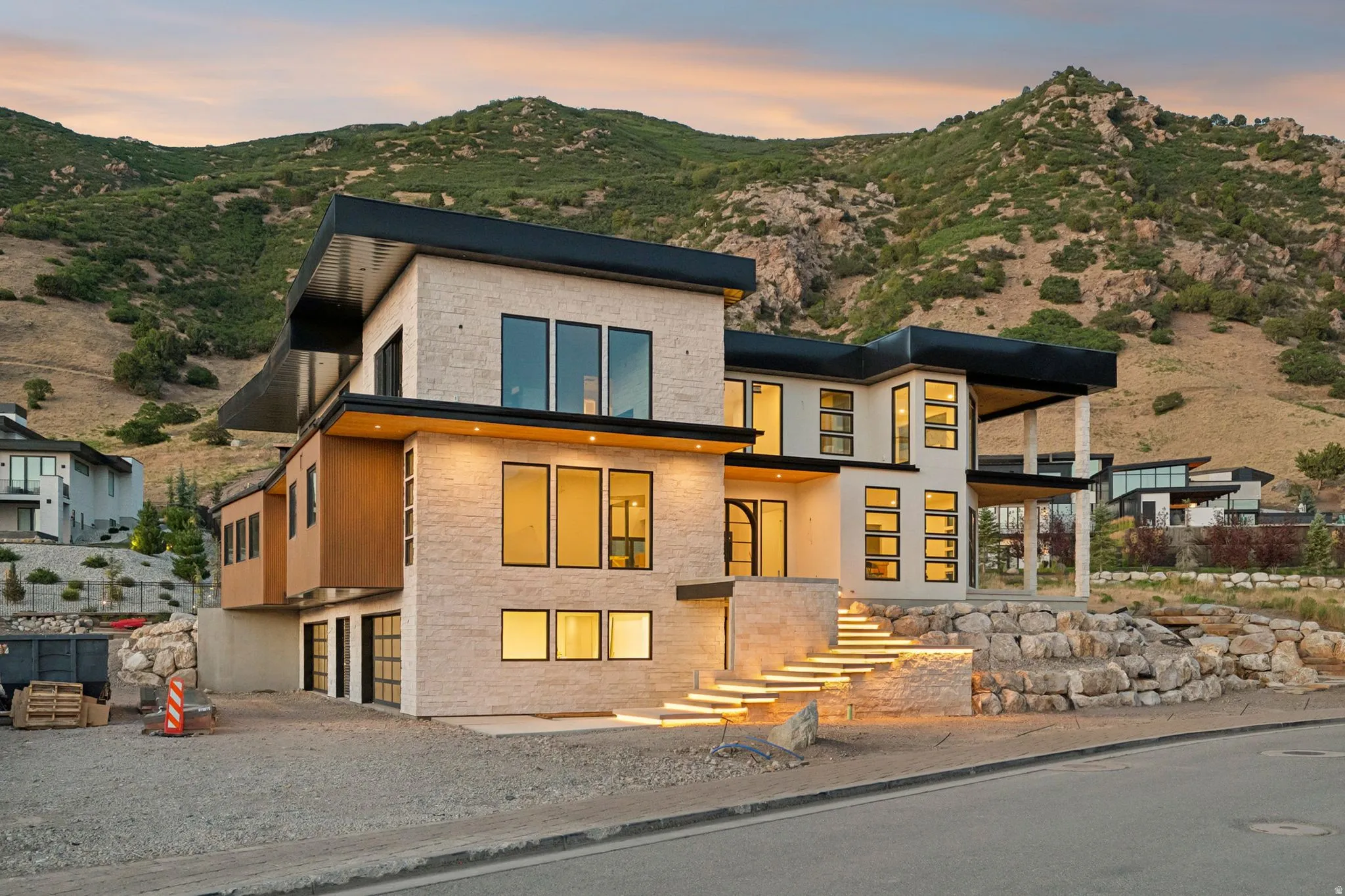 Modern architectural design featuring a stone exterior, flat roofline, integrated lighting on exterior steps, expansive windows, and a mountain backdrop