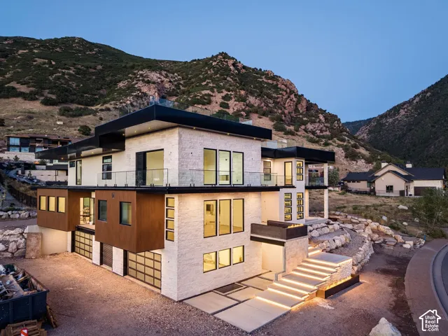Multi-level contemporary exterior featuring stone and wood-finish siding, extensive glass paneling, multiple balconies with glass railings, and an illuminated modern entry staircase