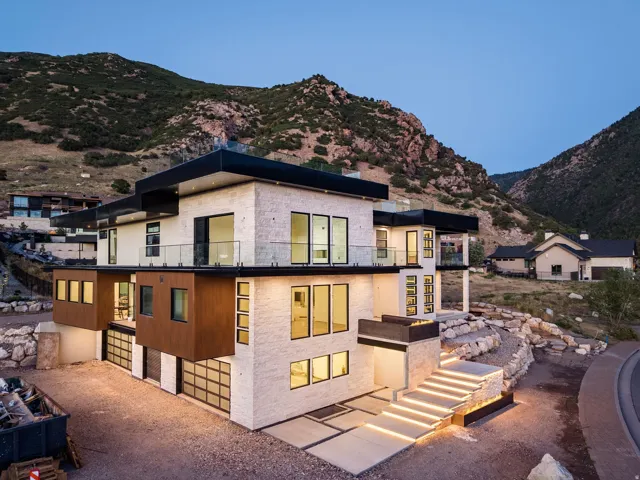 Multi-level contemporary exterior featuring stone and wood-finish siding, extensive glass paneling, multiple balconies with glass railings, and an illuminated modern entry staircase
