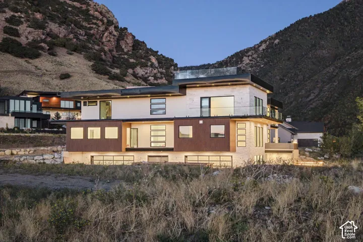 Contemporary multi-level exterior featuring stacked stone masonry, horizontal metal panel siding, a rooftop terrace with glass railing, and multiple glass-paneled garage doors set against a mountainous backdrop