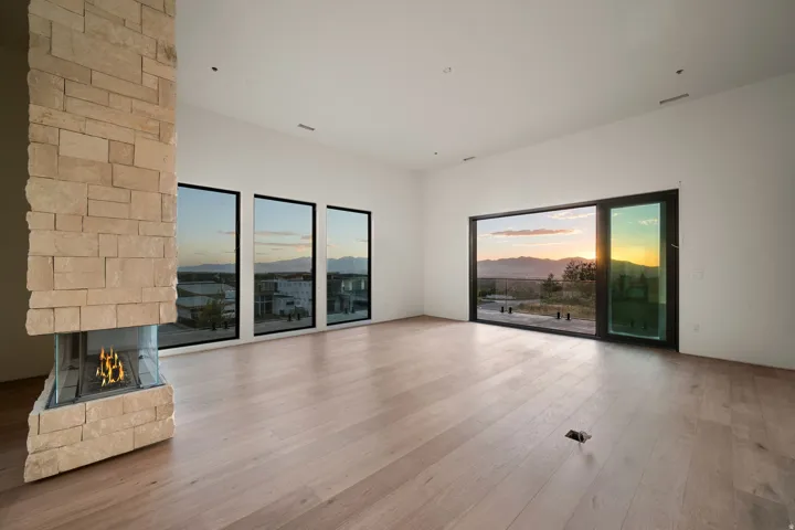 Stone-clad fireplace with a three-sided glass enclosure, wide-plank wood-finish flooring, multiple floor-to-ceiling windows, and a large sliding glass door
