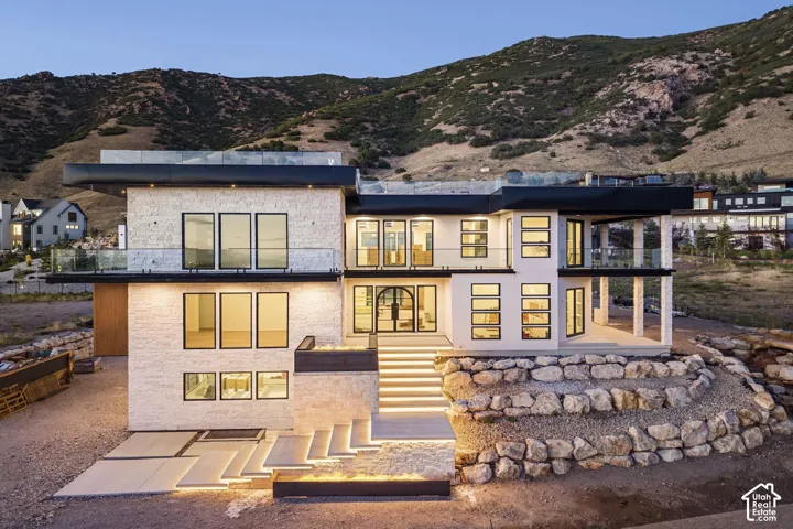 Contemporary residence featuring a multi-level stone facade, expansive glass railings, floor-to-ceiling windows, integrated stair lighting, and a mountain backdrop