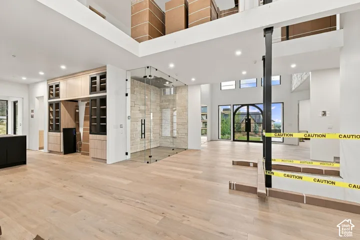 Open-concept interior featuring light wood-finish flooring, soaring ceilings, floor-to-ceiling glass-enclosed stone shower, custom cabinetry with integrated shelving, and an arched glass front entryway