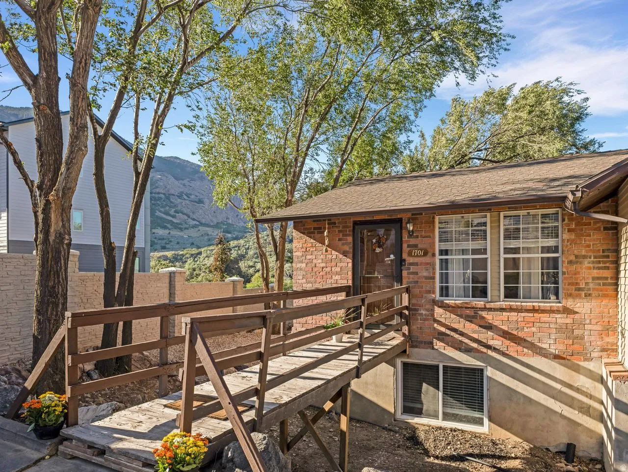 Brick exterior with a brown shingle roof, featuring a wood-framed entry ramp, mature trees, and a mountain backdrop
