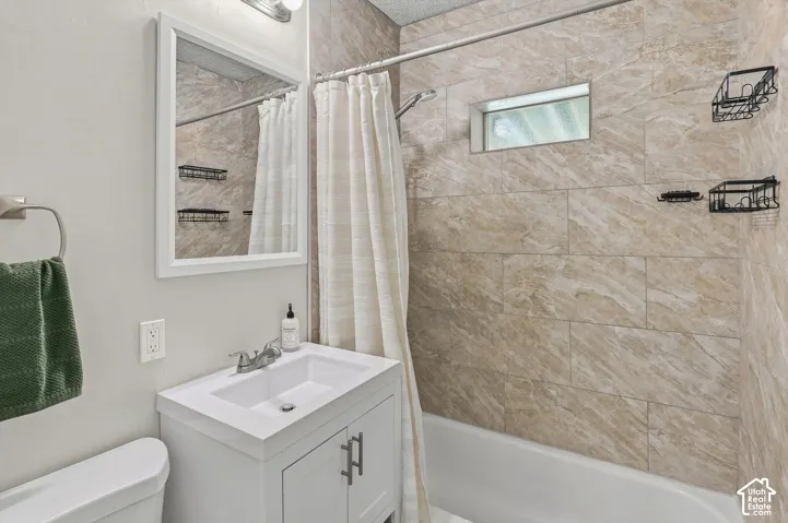 White vanity with an integrated sink and brushed nickel hardware, framed mirror, wall-mounted light fixture, and stone-look tiled shower surround with an inset window