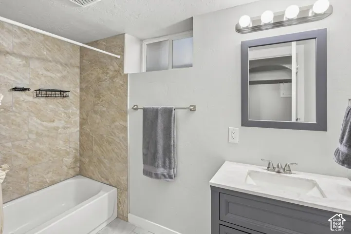 Bathroom featuring a built-in bathtub with tiled surround, a vanity with a white countertop and integrated sink, a framed mirror, and a wall-mounted light fixture