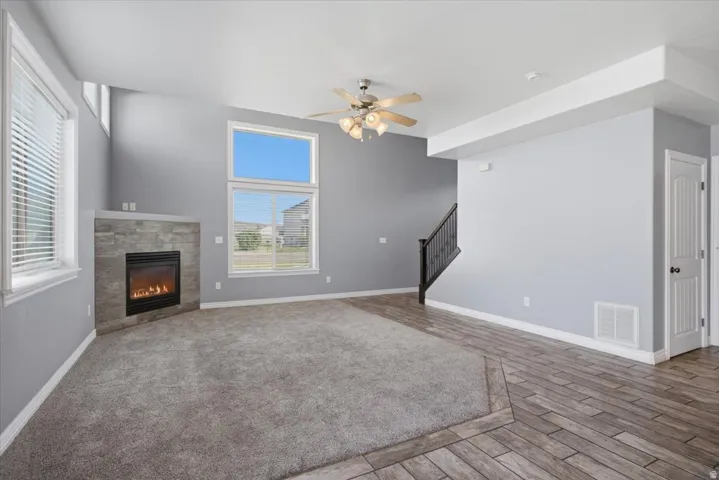 Vaulted ceiling with a ceiling fan, stone-clad fireplace, large windows, and mixed flooring of carpet and wood-finish tile