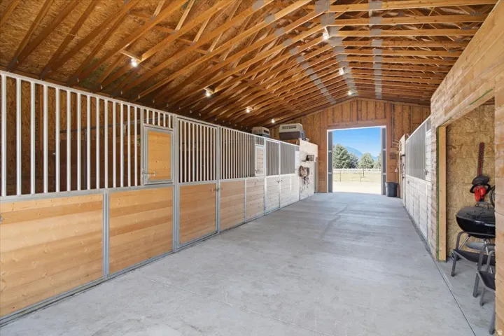 Exposed wood-beam ceiling, multiple stalls with wood-finish panels, concrete flooring, and an open doorway leading to an outdoor area