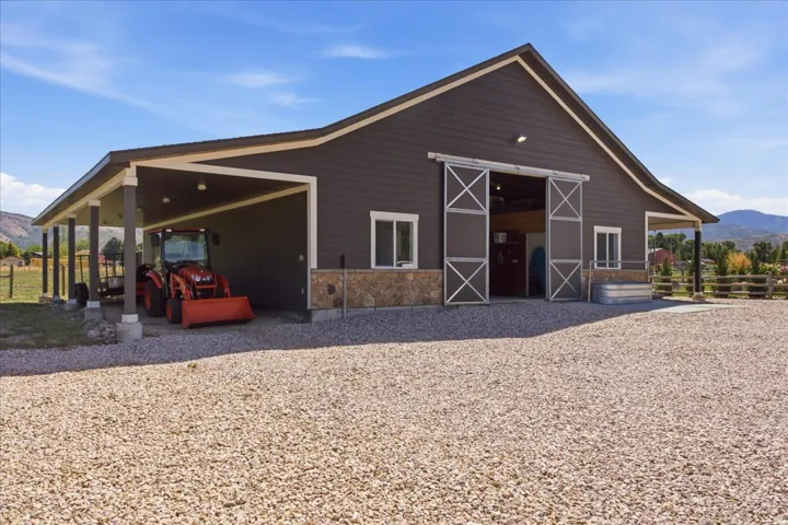 Barn-style building featuring dark siding, stone accents, sliding barn doors, a covered carport, and a gravel driveway