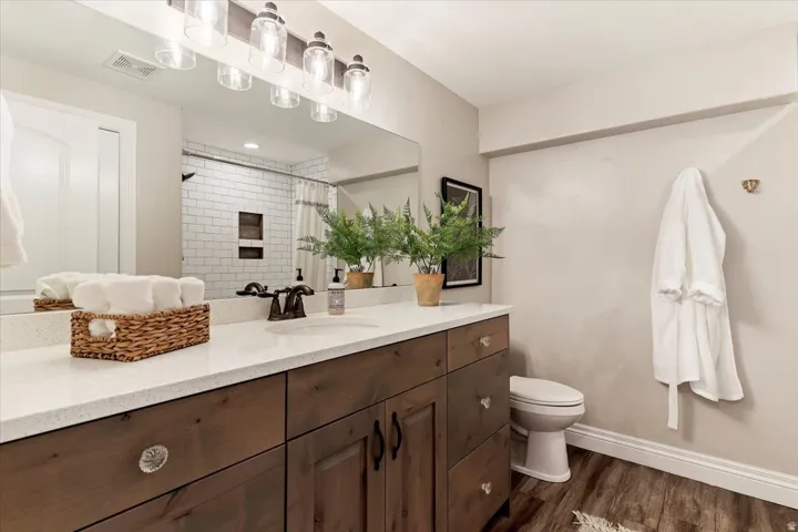 Wood-finish flooring, a wood-finish vanity with crystal knobs, a white countertop, and a large mirror with overhead lighting