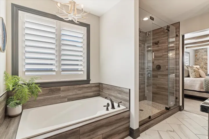 Built-in soaking tub with wood-finish tile surround, walk-in shower with glass enclosure, white plantation shutters, and a decorative antler-inspired chandelier