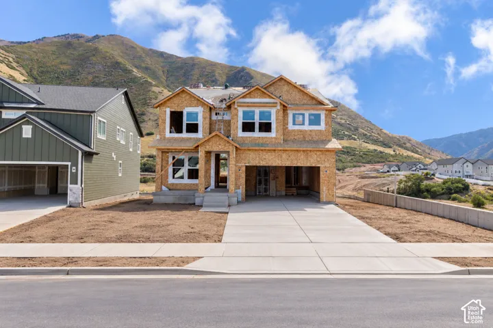 Two-story residential structure under construction featuring oriented strand board exterior, multi-gabled roofline, concrete driveway, and mountain backdrop