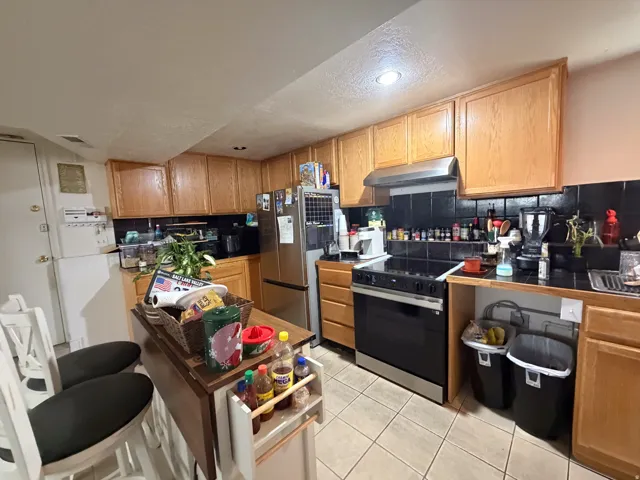 Wood-finish cabinetry with full overlay doors, stainless steel refrigerator, stainless steel range with overhead hood, black tile backsplash, and light tile flooring