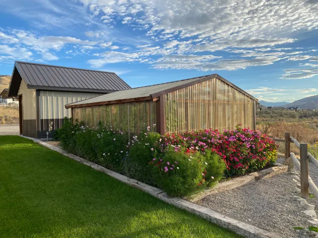 Two-tone corrugated metal outbuilding with a dark metal roof, an adjacent greenhouse with a wood frame, and a vibrant flower bed