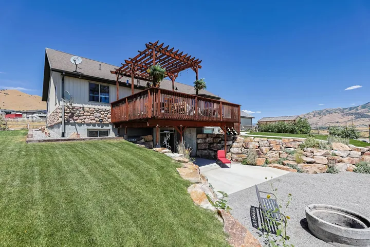 Raised wood deck with pergola, stone retaining wall, large backyard lawn, and a concrete patio area