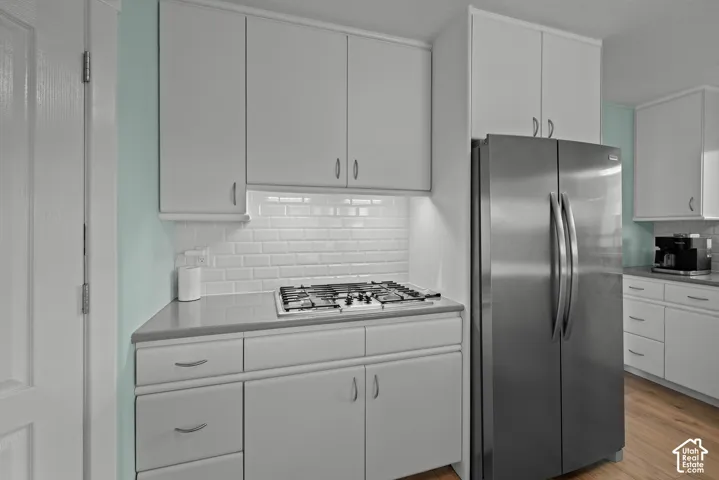 White cabinetry, subway tile backsplash, stainless steel refrigerator, built-in gas cooktop, and wood-finish flooring