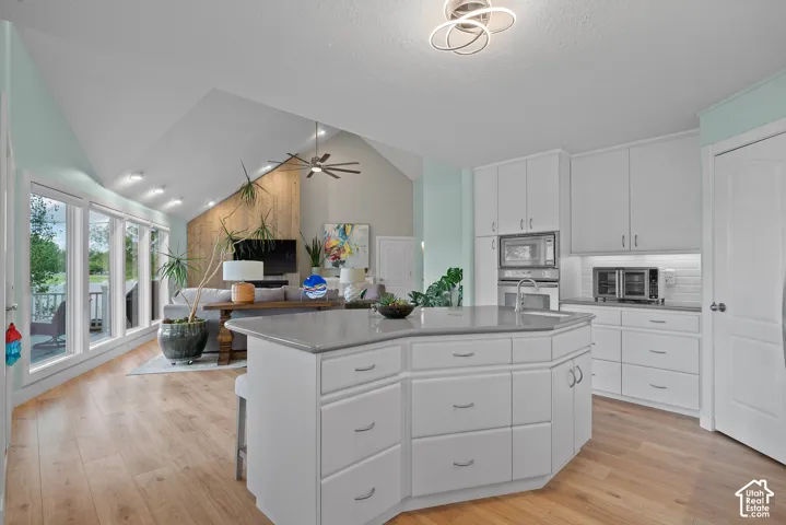 Open-concept kitchen with white cabinetry and an island with a sink, extending into a living area with vaulted ceilings, wood-finish flooring, and a wall of windows
