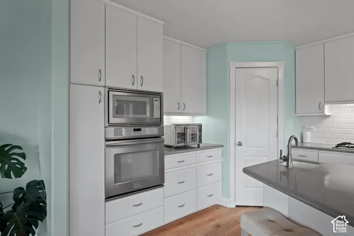Stainless steel built-in microwave and oven, white cabinetry with hardware, wood-finish flooring, white subway tile backsplash, and a modern pull-down faucet
