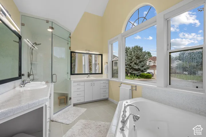 Vaulted ceiling with an arched window, dual vanity with undermount sinks, glass-enclosed shower, built-in soaking tub with deck-mounted fixtures, and light-toned floor tile