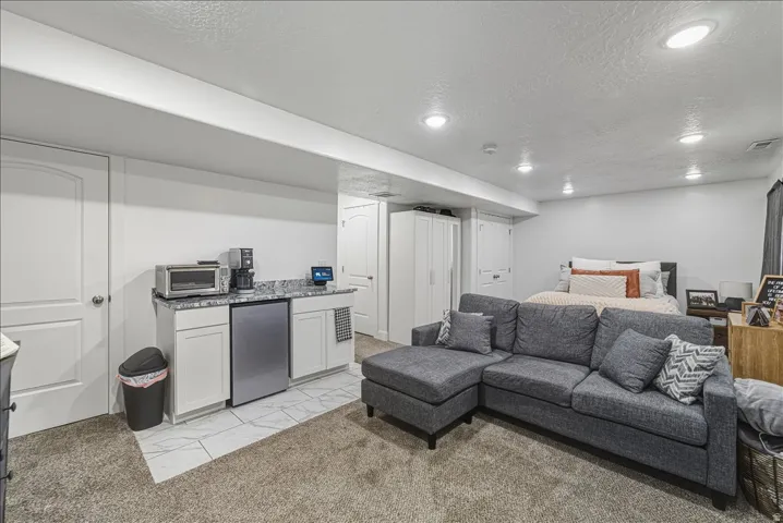 Open-concept living space with recessed lighting, integrated kitchenette featuring white cabinetry, stone-finish countertop, and stainless steel compact refrigerator, and durable carpet flooring