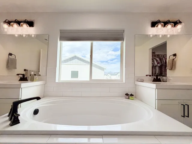 Built-in soaking tub with black fixture, flanked by dual vanities with integrated sinks, and subway tile backsplash