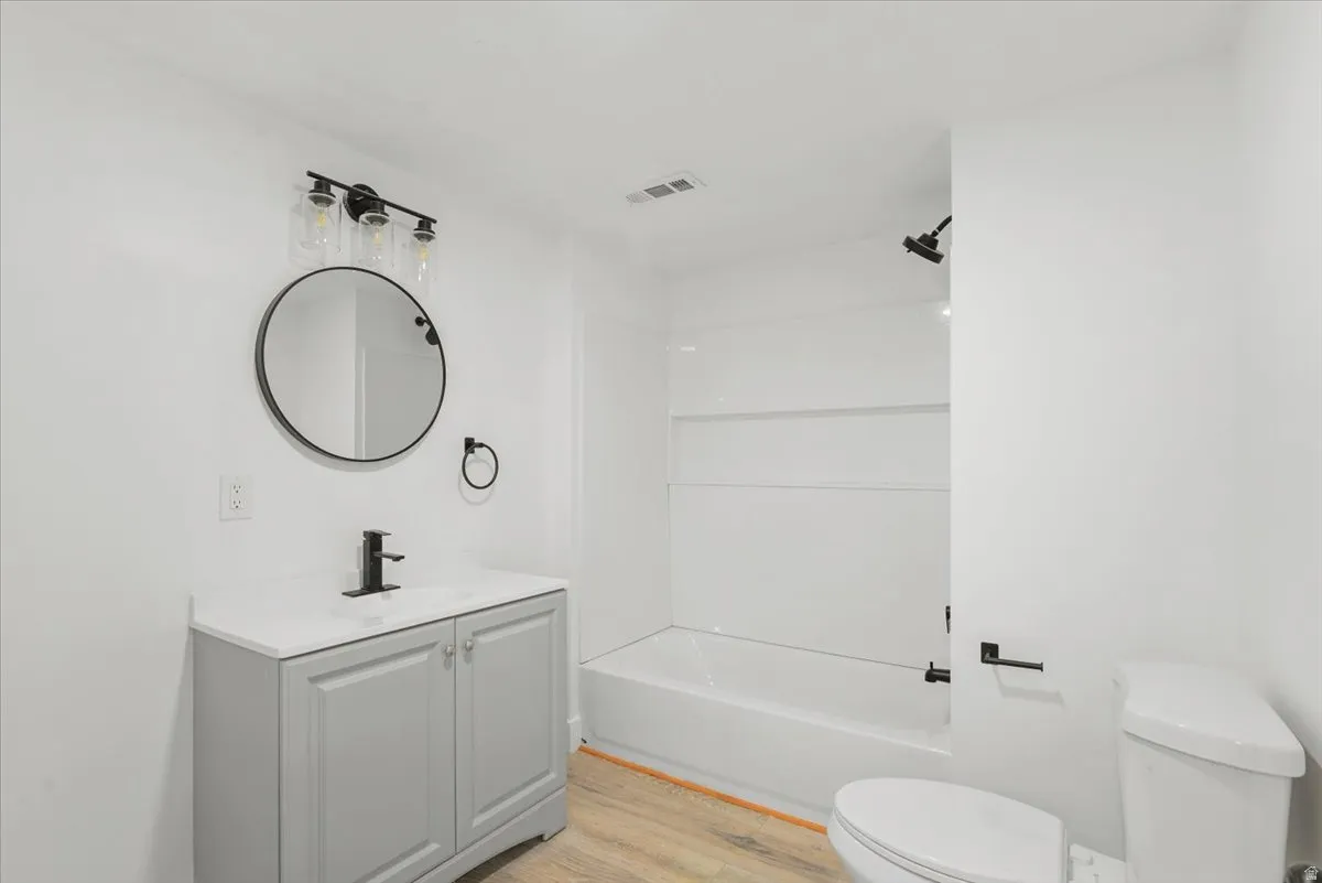 Light gray vanity with white countertop, black fixtures, round mirror, and wood-finish flooring