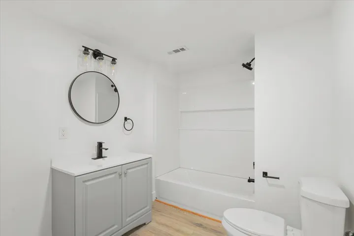 Light gray vanity with white countertop, black fixtures, round mirror, and wood-finish flooring