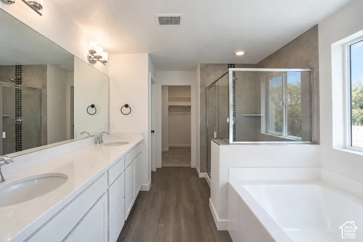 Dual vanity with white cabinetry, wood-finish flooring, built-in soaking tub, and glass-enclosed shower with tile surround
