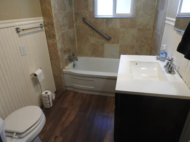 Updated bathroom featuring a soaking tub with grab bar, white quartz countertop with integrated sink, wood-finish flooring, tile surround, and dual windows providing natural light