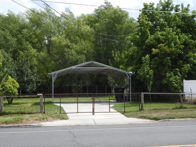 Paved driveway leading to a metal carport structure, surrounded by a wire fence with decorative posts and a gate
