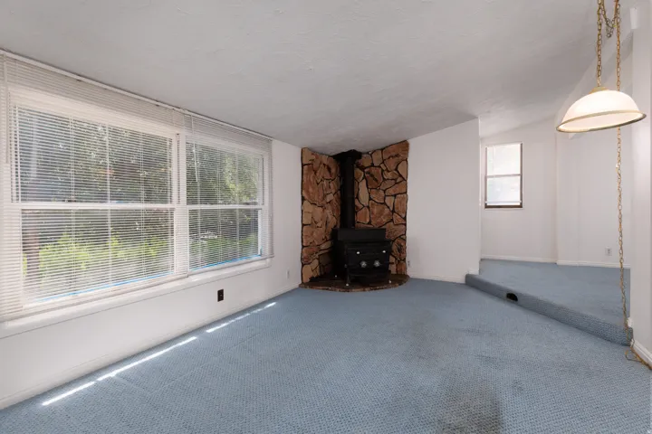 Living area featuring a stone accent wall with a built-in wood-burning stove, light blue carpeting, and dual windows with horizontal blinds