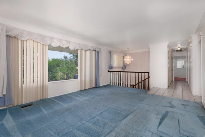 Spacious carpeted living area featuring a large window with vertical blinds, a decorative valance, and crown molding
