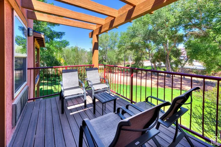 Spacious balcony featuring wood-finish decking, an open pergola, and a dark railing overlooking green space