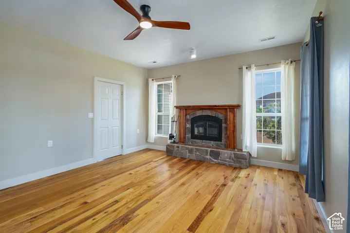 Spacious room featuring wood-finish flooring, a stone and wood fireplace, two windows, a white paneled door, and a ceiling fan with integrated lighting