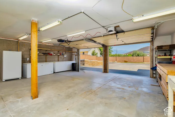 Concrete flooring with exposed wood support beam, fluorescent lighting, and open garage door revealing a paved area and distant mountains