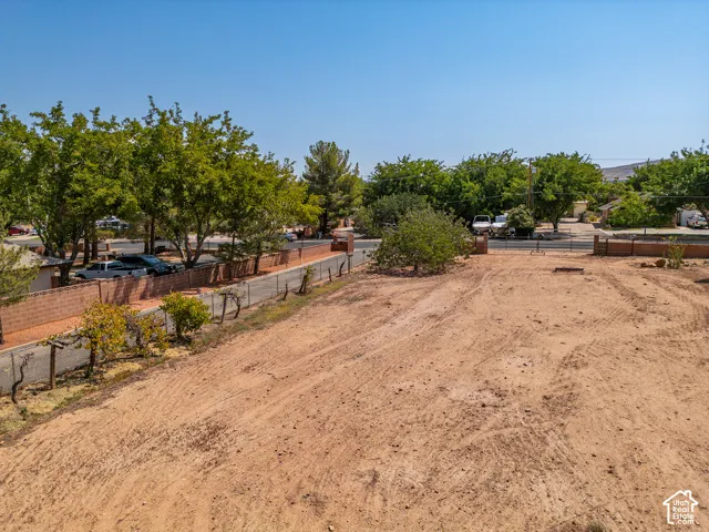 Expansive undeveloped lot with clear ground, bordered by a low brick wall and fencing, featuring mature trees and grape vines.