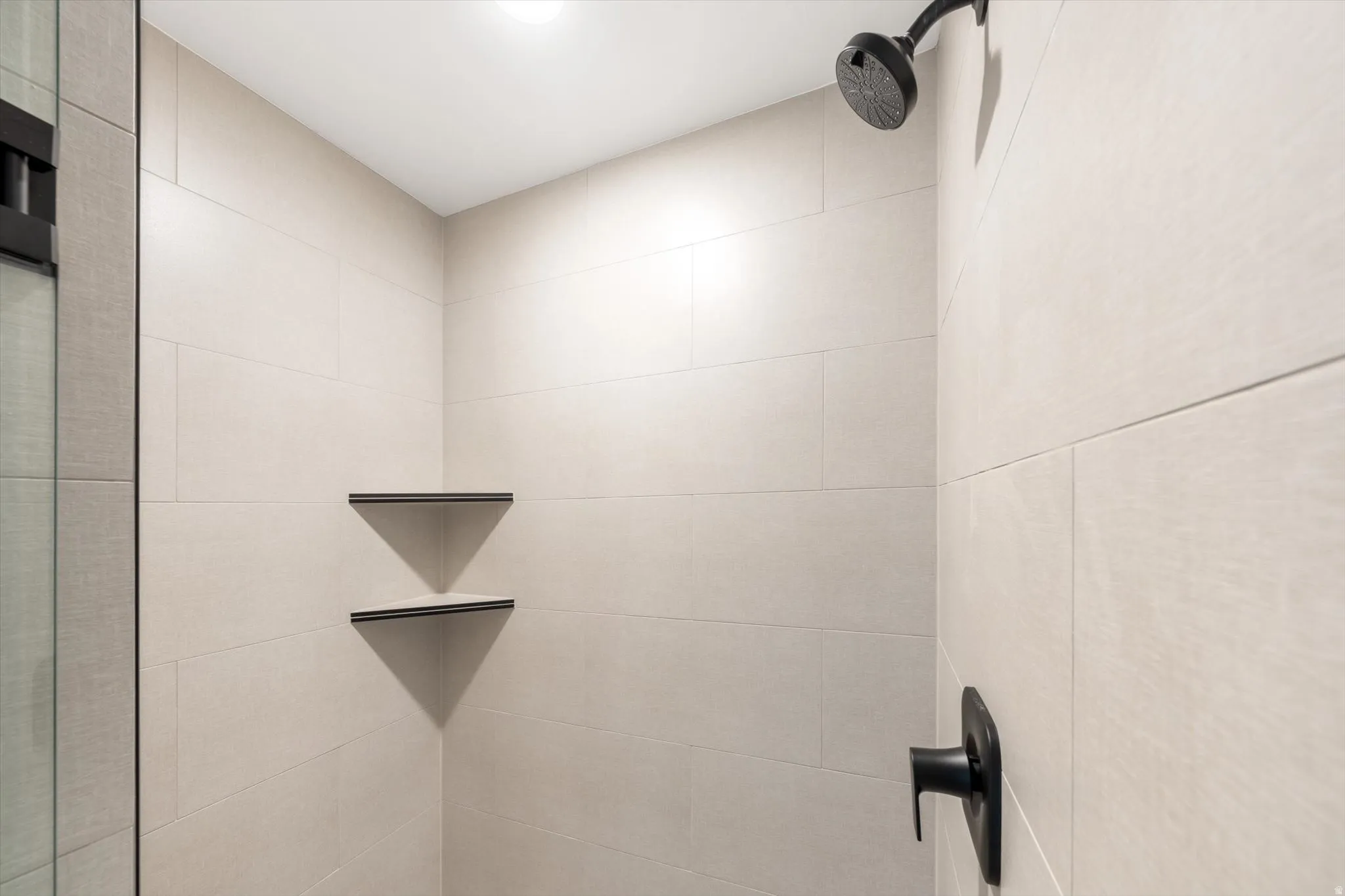 Shower stall featuring light-toned tile walls, matte black shower head, matte black shower control, and two built-in corner shelves