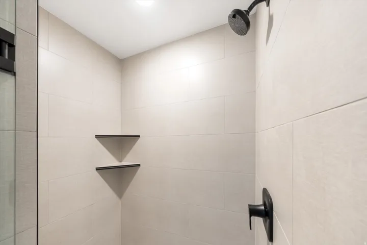Shower stall featuring light-toned tile walls, matte black shower head, matte black shower control, and two built-in corner shelves