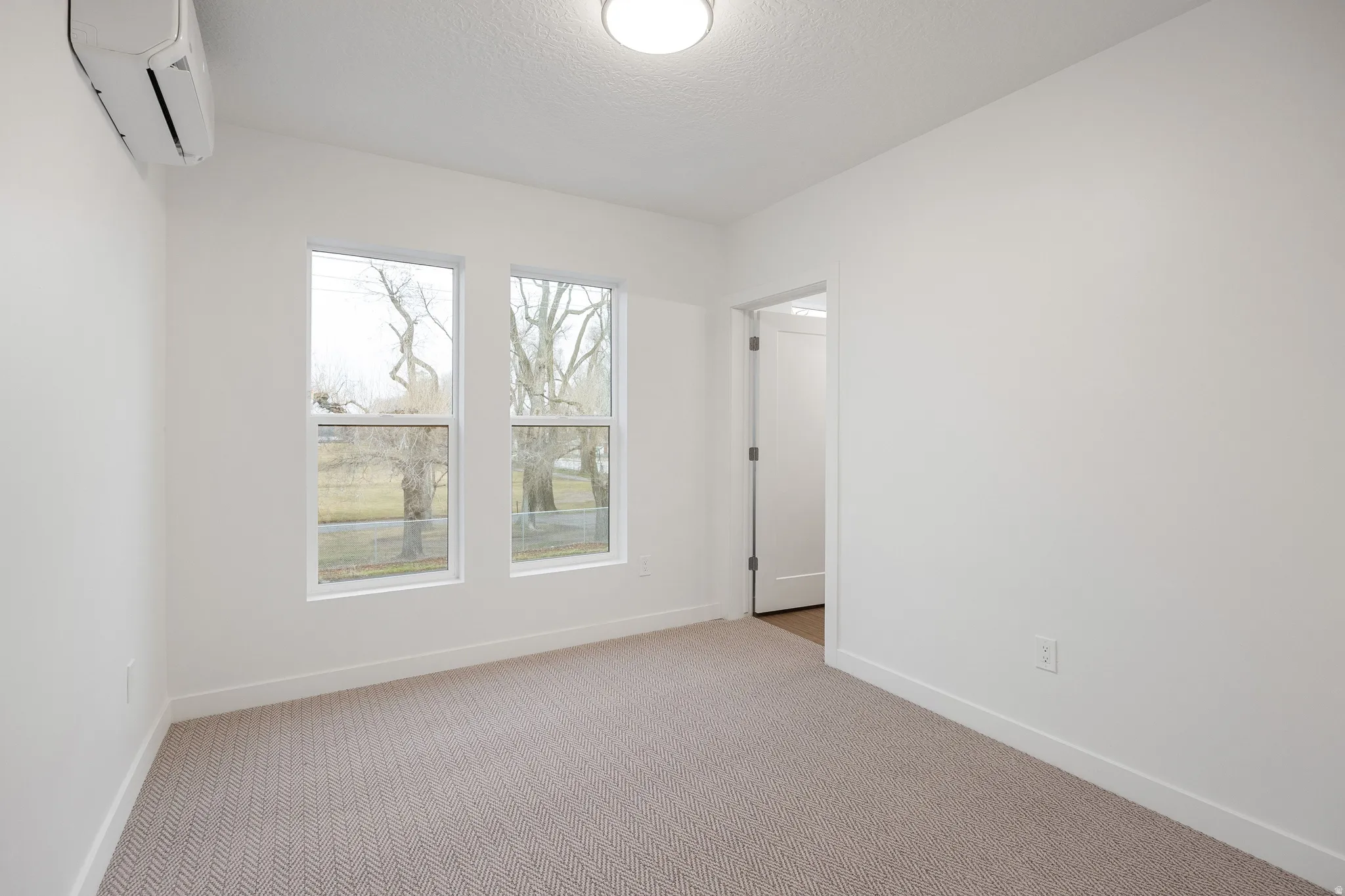 Crisp white walls, two single-hung windows, light-colored patterned carpeting, a white overhead flush-mount light fixture, and a wall-mounted mini-split unit