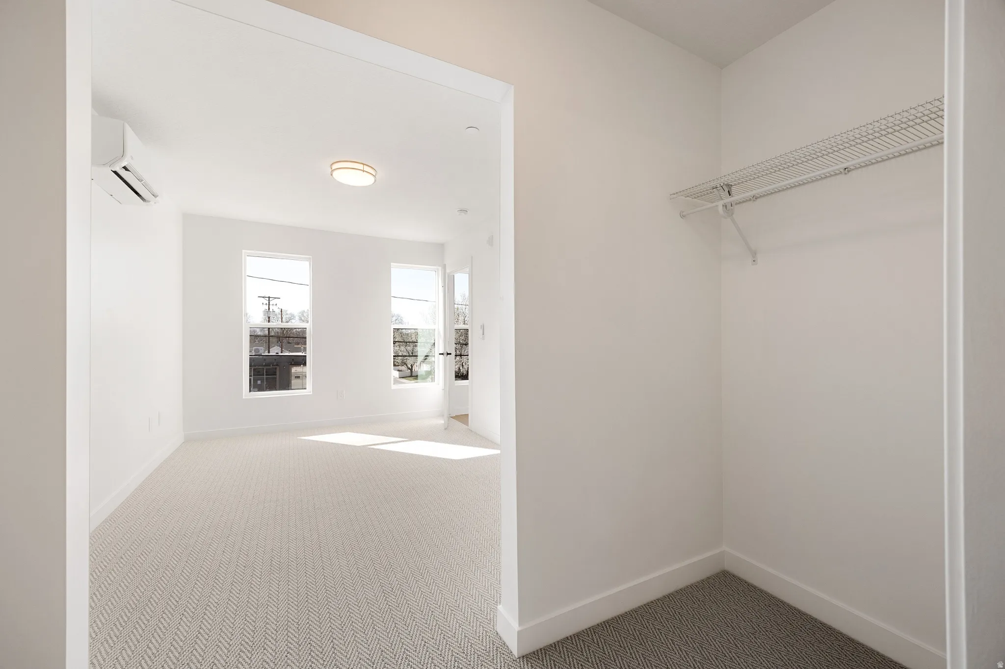 Room featuring a textured carpet, two windows, a ceiling-mounted light fixture, and a wall-mounted climate control unit