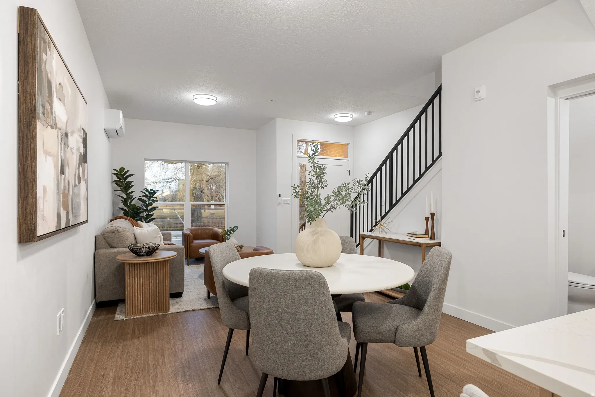 Open-concept living and dining area with wood-finish flooring, a white round dining table, and a contemporary black metal staircase railing