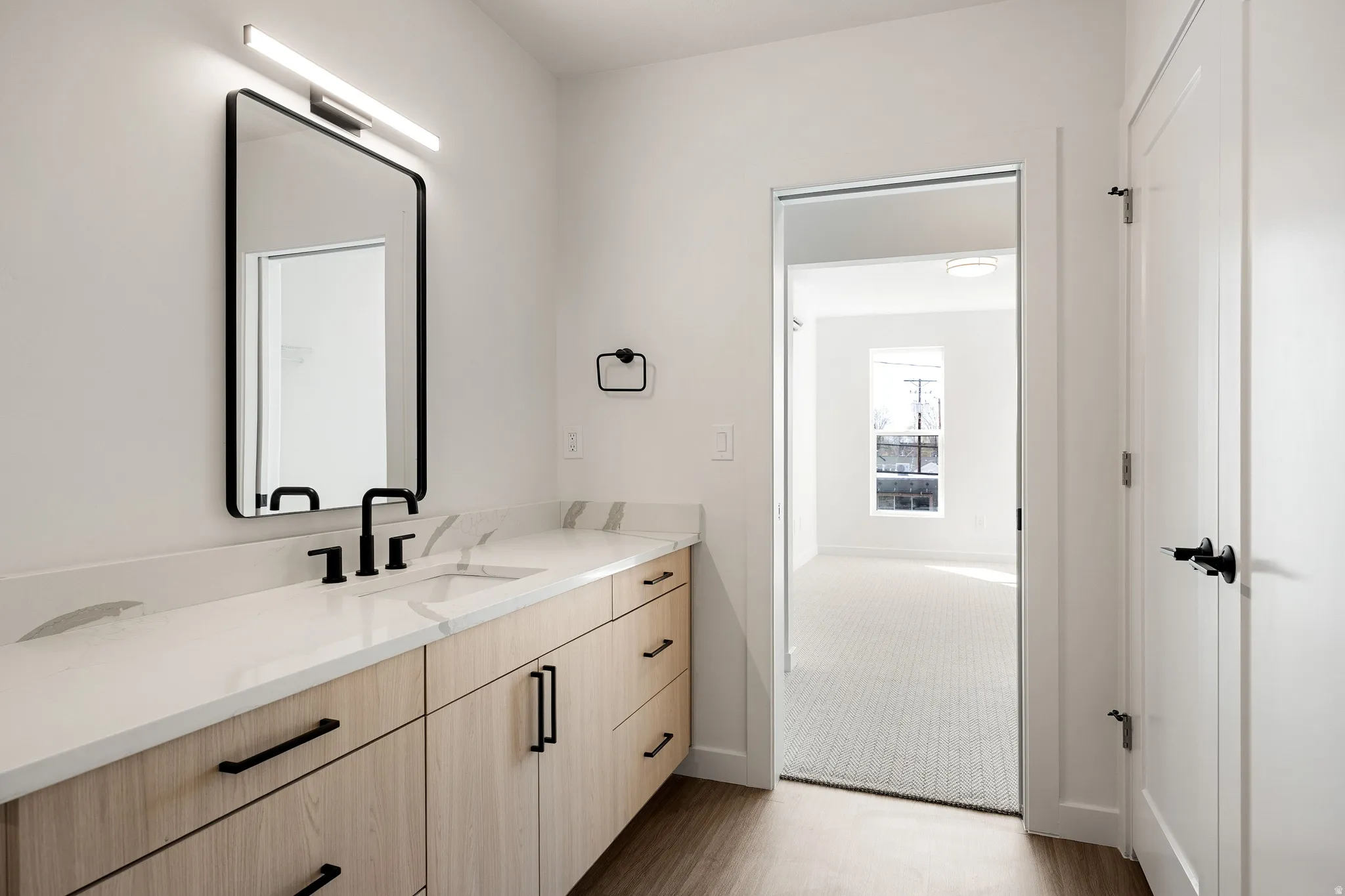 White countertop with integrated sink, black fixtures, wood-finish cabinetry, and contemporary mirror with overhead lighting