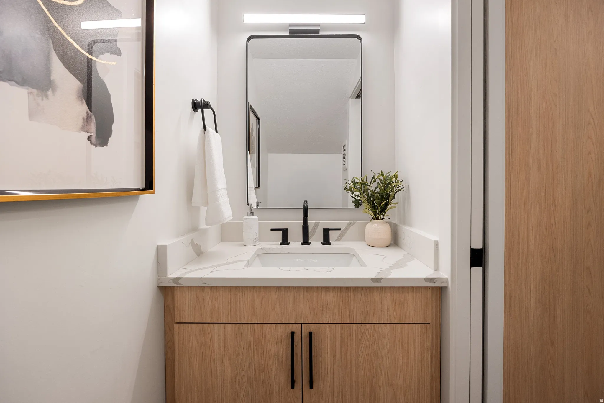 Light-toned wood vanity with integrated sink and matte black faucet, framed mirror with horizontal vanity light, and white countertop with veining