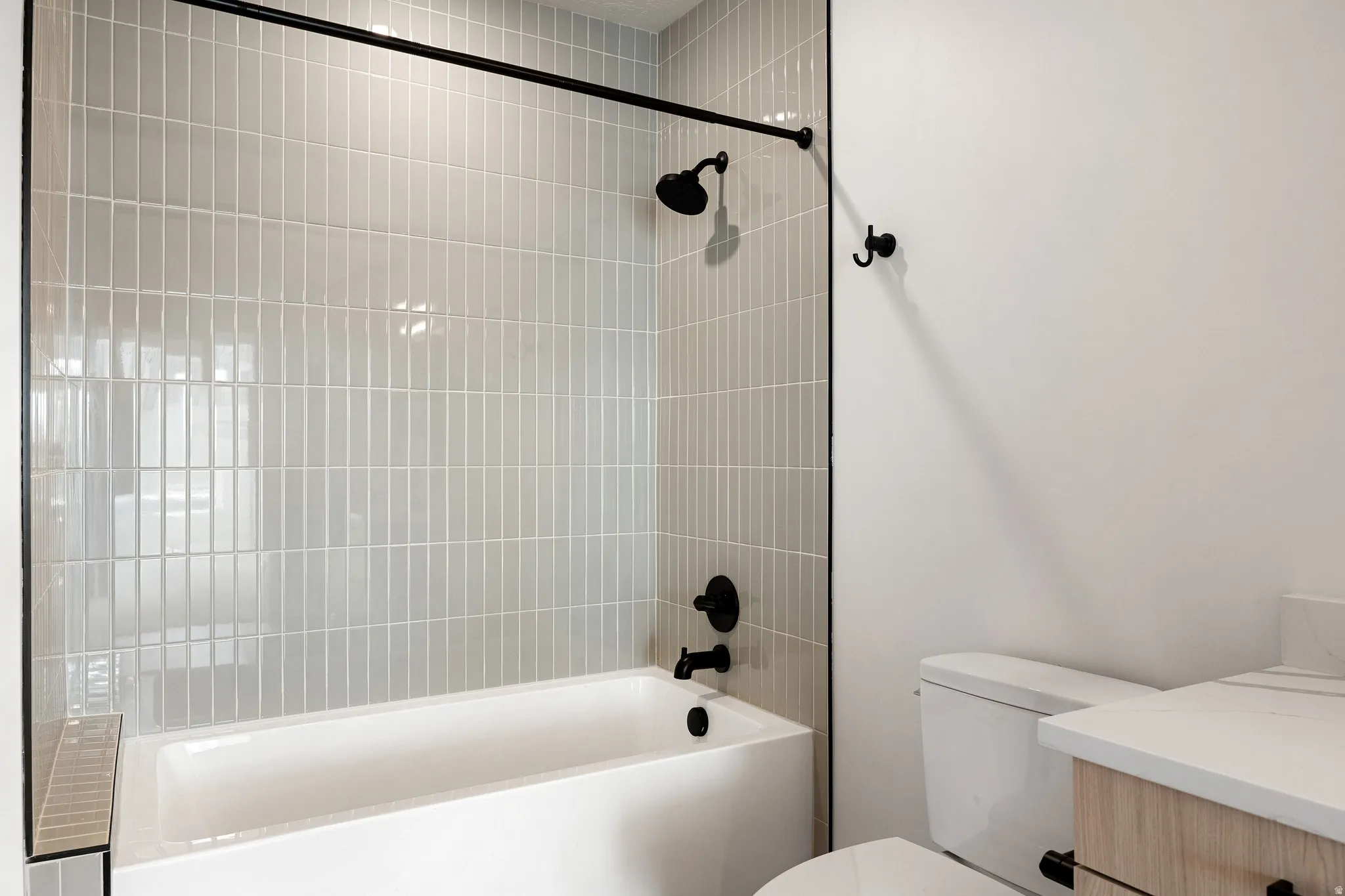Bathtub and shower combination with vertical stacked tile surround, matte black shower fixtures, and a light wood-finish vanity with a light countertop