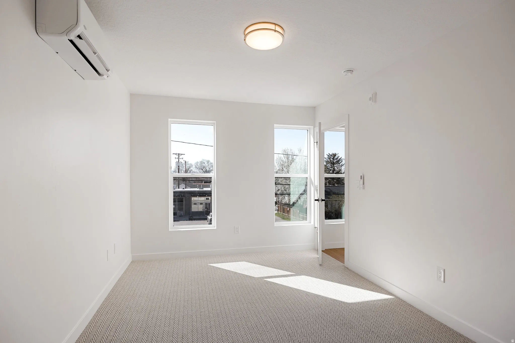 White walls, two windows, wall-mounted air conditioning unit, ceiling-mounted light fixture, and light-colored carpet